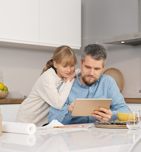 Mies selaa tablettia pöydän ääressä yhdessä lapsen kanssa.